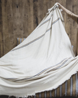 A person spreads the Libeco Home Auburn Coverlet, a large cream-colored towel with navy and bronze stripes, over a bed with striped bedding, set against a wooden wall background.