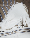 The Libeco Home Auburn Coverlet, in cream with navy and bronze stripes, is draped over a bed, coordinating with striped pillows and complementing the wooden wall behind them.