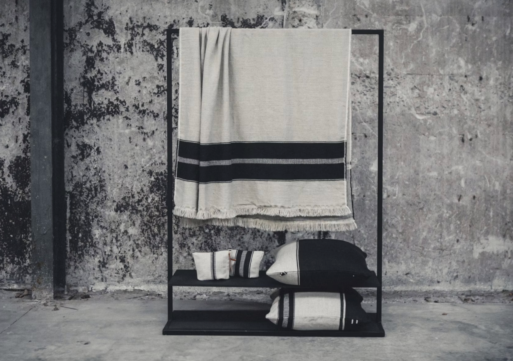 A minimalist black metal rack against a weathered concrete wall displays the Libeco Home Marshall Throw—a beige blanket with black stripes and flax fringe—paired with matching linen and wool towels and pillows for a timeless look.