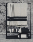 A minimalist black metal rack against a weathered concrete wall displays the Libeco Home Marshall Throw—a beige blanket with black stripes and flax fringe—paired with matching linen and wool towels and pillows for a timeless look.