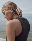 A blonde woman wearing The Not So Basic Tank by Sold Out NYC stands by the water, touching her neck and looking down thoughtfully against a calm, cloudy sky background.