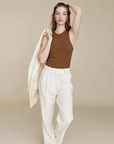 A woman with wavy hair and a confident expression stands against a beige background, wearing Sold Out NYC's The Not So Basic Tank in brown and high-waisted cream trousers, holding a cream blazer over her shoulder.