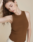 A woman with wet, wavy hair and gold earrings poses calmly against a beige background, wearing Sold Out NYC’s The Not So Basic Tank in brown with cream pants. She looks down, one hand in her hair, showcasing the ribbed cotton design.