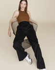 A woman with wet hair sits in a modern wire chair, wearing Sold Out NYC’s The Not So Basic Tank in brown, paired with high-waisted black pants and white Adidas sneakers, against a plain beige background.