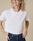 A woman in Sold Out NYC's The Cropped Perfect Tee, plain white Peruvian cotton, paired with dark blue high-waisted jeans and large sunglasses, smiles with tousled hair against a beige background.