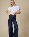 Wearing The Cropped Perfect Tee by Sold Out NYC and dark blue wide-leg jeans, a blonde-haired person stands before a beige backdrop, embodying the relaxed ease of a summer breeze.