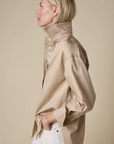 A person with blond hair stands in profile against a beige background, wearing Sold Out NYC’s The Everything Shirt—an oversized beige button-up with the collar popped—and white pants, with one hand in their pocket.