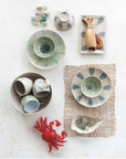 A flat lay features the Creative Co-op Low Bowl Blue Green with other ceramic dishes, cups, and bowls in blue-green patterns, a woven placemat, glass marbles, a wooden seal figurine, a red crab figurine, and a shell-shaped dish on white.