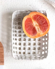 A halved red grapefruit rests on a postcard-sized Creative Co-op Square Lattice Dish atop a light surface. In the lower left, part of a wooden board with assorted fruits is visible.