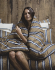 Wrapped in the Salem Duvet by Libeco Home, a woman sits on a bed with matching striped linen, gazing at the camera. The 100% linen fabric and wooden wall background create a warm, inviting atmosphere.