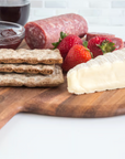 A beautifully arranged Provencale Acacia Board by Fox Run Brands holds sliced salami, crackers, brie cheese, strawberries, jam, and a glass of red wine for the perfect charcuterie spread.