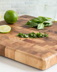 The Square Chef's Board by Fox Run Brands holds chopped herbs, a knife, basil leaves, a lemon, a whole and sliced lime. In the background are a bowl of greens and two glasses of water with lime.