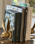 The Fox Run Brands Rabbit Bookend Set holds four books—"CHAMPAGNE," "The Italian Art of Shoemaking," "Carolina Herrera," and "Stage Set 50 Years of Style"—on a round wooden table.