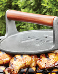 The Fox Run Brands Cast Iron Grill Press, featuring a wooden handle, rests atop grilled chicken on an outdoor barbecue and doubles as a bacon press, with green foliage visible in the background.