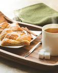 The Fox Run Brands Acacia Serving Tray holds a white mug of steaming tea, two sugar cubes, a plate with croissants and cinnamon sticks, with a folded green towel in the background.