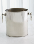 The Winston Ice Bucket L by Global Views, crafted from stainless steel with a fluted design and round side handles, rests on a light surface with a white curtain backdrop.