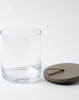 The Rustic Canister MD by Global Views features a clear cylindrical glass jar and a round wooden lid with a leather loop handle, shown on a white background—ideal for rustic kitchen storage or as stylish modern canisters.