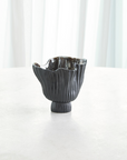 The Folha Footed Vase S by Global Views is a dark ceramic piece with a wavy, fluted rim and vertical ridges, hand-painted in metallic tones by Portuguese artisans. It sits on a round surface with sheer white curtains in the background.