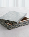 The Interwoven Box LG by Global Views features a geometric grey pattern and is shown partially open on a light surface with a soft white curtain in the background.