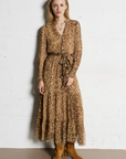 A woman with light hair stands against a white wall wearing the SUNCOO Cosmos Dress, a loose-fit, long brown leopard print style with a tie waist, paired with tan suede ankle boots and layered necklaces. Dress is 100% viscose.