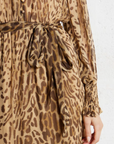 Close-up of a person wearing the SUNCOO Cosmos Dress, a loose-fit brown leopard print dress with a matching fabric belt at the waist. Made from 100% viscose, it has long sleeves with smocked cuffs and the person's hand is at their side.