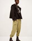 A person with short hair poses in the BA&SH Jacket Kove over a dark hoodie with a white “G,” light olive-green drawstring pants, and black boots, standing against a plain light background.