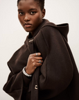A woman with short hair wears the BA&SH Jacket Kove, a dark brown, loose-fitting jacket with a hood, metallic accents, and wide collar. She holds it closed, showcasing her rings and bracelet as she looks confidently at the camera against a plain background.