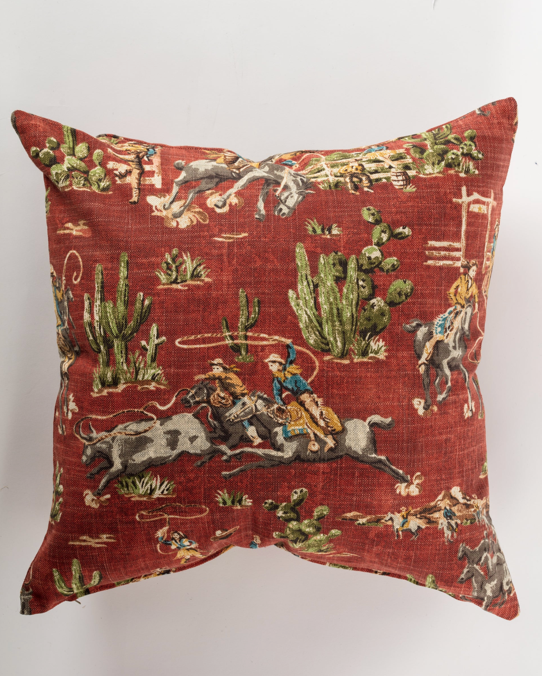 The Pindler Cowboys 26x26 Pillow showcases a red background with Western-themed cowboys riding horses and bulls among green cacti—perfect for adding flair to your Western decor.
