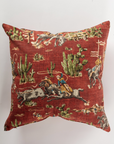 The Pindler Cowboys 26x26 Pillow showcases a red background with Western-themed cowboys riding horses and bulls among green cacti—perfect for adding flair to your Western decor.