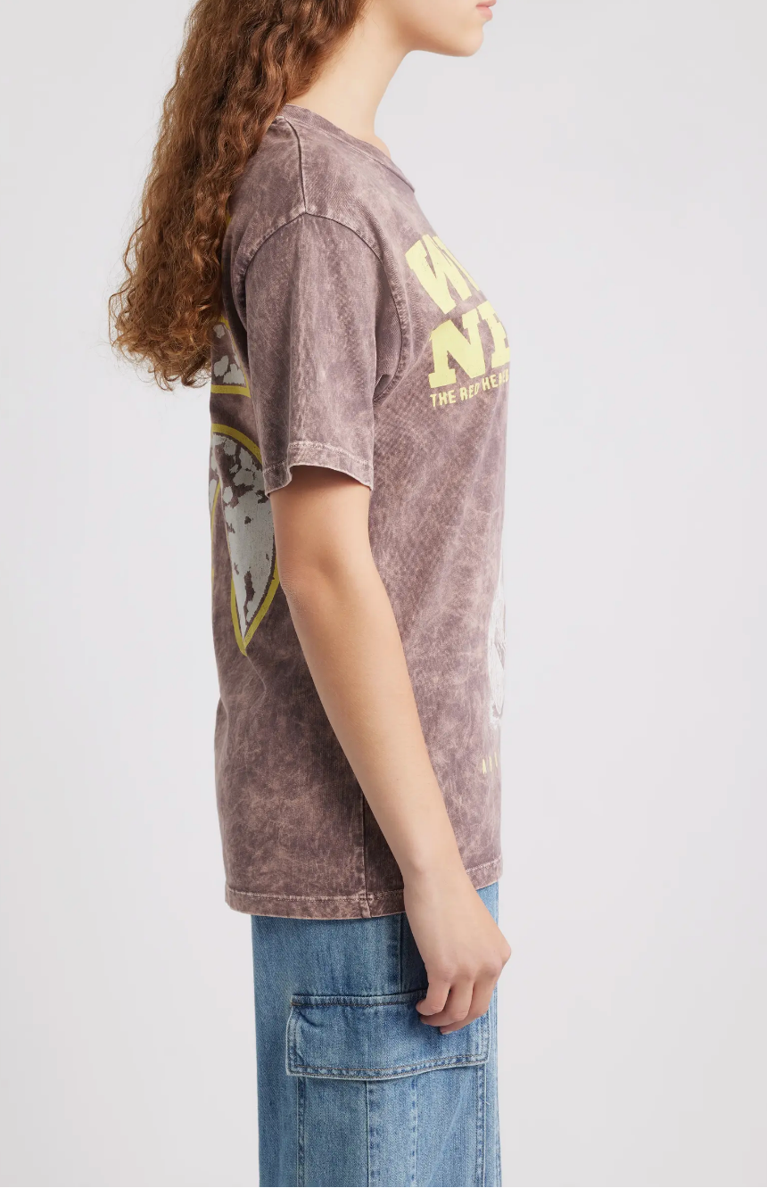 A woman with long curly hair stands in profile against a plain white background, wearing the DayDreamer Willie Nelson Red Headed Stranger Tee and blue jeans with large pockets.