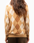 A person with long, wavy brown hair is shown from the back wearing the Brazeau Tricot Argyle Cardigan in brown, beige, and cream, paired with a brown skirt, against a plain white background.