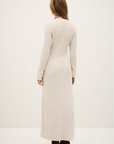 A woman with light brown hair wears the BA&SH Dress Anisa, a long, cream ribbed knit dress with long sleeves, made from a cozy polyester blend. She pairs it with brown heeled boots against a plain light background.