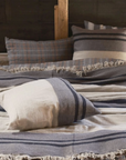 A cozy bed features Libeco Home’s Black House Stripe Pillow, styled with 25x25 inserts. The striped design in gray, cream, and navy rests against a wooden headboard, evoking rustic warmth and comfort.