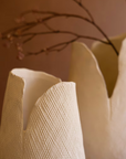 The Bloom Flower Vase S by Kalalou, Inc features a white textured finish with jagged, organic edges. Against a soft brown background, it holds thin branches with small dried flowers, adding a natural, minimalist charm to any space.