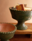 Two Green Terra Clay Compote bowls by Kalalou, Inc sit on wood; one holds colorful cheese blocks, the other is empty. A blurred, warm-toned background enhances the inviting decorative scene.