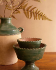 The Green Terra Clay Compote by Kalalou, Inc is stacked on a wooden surface, paired with a beige and green vase holding dried ferns for a rustic, earthy still-life display.