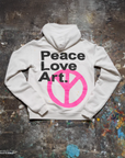 The Made Worn Peace Love Oversized Hoodie rests on a paint-splattered surface. Its back features bold black "Peace Love Art." text above a large pink peace symbol, making it a cozy winter pick for creative spirits.