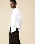 A person stands sideways against a beige background, confidently wearing Sold Out NYC's The Everything Shirt—an oversized, 100% cotton white button-down—paired with wide-leg black pants, hands in pockets.