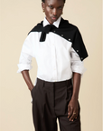 A person stands with one hand in their pocket, wearing Sold Out NYC’s The Everything Shirt— a tailored 100% cotton white button-up—paired with dark brown trousers and a black sweater draped over their shoulders against a beige background.