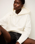 A person with short hair wears the BA&SH Sweatshirt Didi—a white, washable polyester cotton hoodie—styled with pins, black leather shorts, and a chain necklace, sitting against a white background.