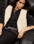 A woman with long dark hair reclines on a black leather couch, wearing the BA&SH Jacket Celton over a black button-up shirt and black jeans, accessorized with a black belt and silver jewelry.