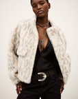 Wearing the BA&SH Coat Saros, a light leopard-print faux fur cropped jacket, over a low-cut black top with silver necklaces, a stylish belt, and black jeans, a person poses confidently against a plain backdrop.
