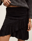A person wears a dark grey textured polyester long-sleeve sweater with the BA&SH Skirt Rino, a black ruffled asymmetrical mini skirt, accessorized with rings and a bracelet. Only the torso and upper legs are visible.