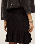 A person wears a textured dark grey polyester sweater and the BA&SH Skirt Rino—black, patterned with a ruffled hem. Gold bracelets and rings adorn their wrists and fingers. Image is shoulder to knee; dry clean recommended.