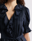 A woman models the DOEN Harriette Dress, featuring navy blue fabric, ruffled collar and front, puffed short sleeves with shirred cuffs, and a waist-cinching belt. Her curly hair frames her shoulders for an elegant look.