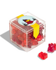 The Faire Christmas Candy Cube features red gummy bear-shaped holiday candies in a clear plastic box, finished with a yellow ribbon and a bear-in-winter-hat sticker—perfect for festive gifting or treating yourself.