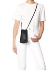 A person in a white t-shirt and jeans models the Jerome Dreyfuss Gordi Mobile, a small black goatskin crossbody bag with a leather strap, ideal as a mobile case, hanging stylishly at hip level against a white background.
