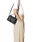 A person in a white t-shirt and beige high-waisted trousers stands in profile, one hand in their pocket, carrying the Jerome Dreyfuss Lucky Mini Bowling Bag—a small black leather bag with a removable shoulder strap.