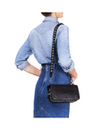 A woman in a light blue denim shirt and skirt stands with her back to the camera, carrying the Jerome Dreyfuss Bandouliere Festival Strap, a black leather shoulder bag with metal studs and a removable strap.