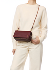 A person in a cream sweater and pants holds the Jerome Dreyfuss Bobi Bag, made of maroon goatskin with an adjustable chain strap, against a white background.